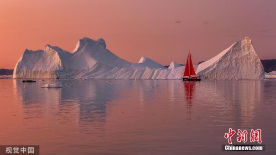 9月5日讯(具体拍摄时间不详)，格陵兰岛迪斯科湾，一艘挂着红帆的游艇在形状各异的冰山之间航行，为宁静的北极夜晚增添了一抹亮色。图片来源：视觉中国